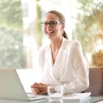 Cheerful businesswoman in glasses working on a laptop, in a bright and modern office setting.